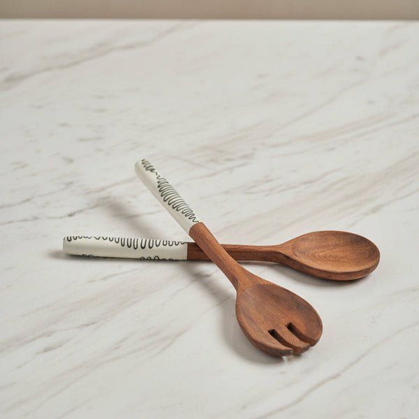 Two wooden serving utensils, one spoon and one fork, with white ceramic handles featuring black wavy line patterns, arranged on a light gray background.