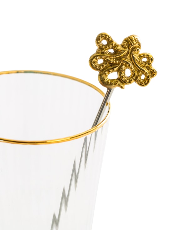 Gold octopus drinks stirrer resting on gold-rimmed glass, white background