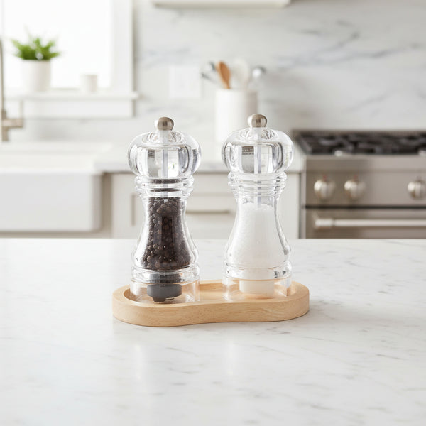 Clear pepper mills with black peppercorns on a wooden base against a white background